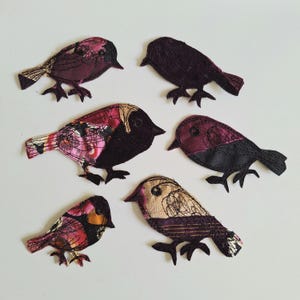 Könnte beinhalten: Sechs Stoffvogel-Applikationen in Burgunder-, Schwarz- und Beigetönen. Jeder Vogel hat ein einzigartiges Design mit unterschiedlichen Mustern und Texturen. Die Vögel sind auf einer weißen Oberfläche angeordnet und zeigen ihre detaillierte Handwerkskunst.