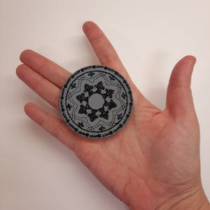 May include: A gray circular coaster with a black embroidered design of a geometric pattern.