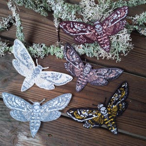 May include: Embroidered moth patches in various colours, including white, grey, purple, and black with yellow accents. The patches are arranged on a wooden surface with greenery.