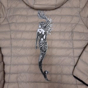 May include: A beige puffer jacket with a black embroidered mermaid design on the back.