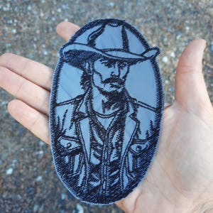 Può includere: Toppa ricamata di una persona che indossa un cappello da cowboy e una giacca. La toppa è di forma ovale e ha uno sfondo blu con ricami neri.