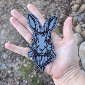 May include: A gray embroidered patch of a rabbit's head with black stitching. The rabbit is holding a bunch of carrots in its mouth.