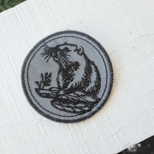 Könnte beinhalten: Ein grauer gestickter Aufnäher mit schwarzem Rand, der ein stilisiertes Bild eines Bibers zeigt.