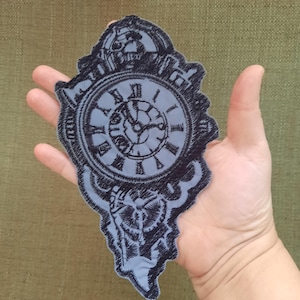 Puede incluir: Un parche bordado gris de un reloj con hilo negro. La esfera del reloj tiene números romanos y las manecillas apuntan a las 12:00.