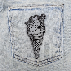 Puede incluir: Un parche bordado en blanco y negro de un cono de helado con tres bolas de helado. El parche está cosido en un bolsillo de mezclilla azul.