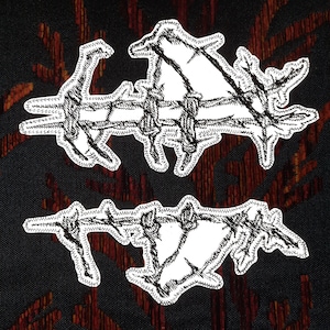 Puede incluir: Dos parches bordados blancos con un diseño de alambre de púas. Los parches están sobre un fondo de tela rojo oscuro y negro.