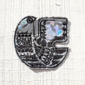 May include: A silver and black fabric patch with a detailed design of a bird with its wings spread. The patch is shaped like a letter C.