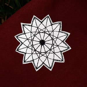 May include: A white star-shaped patch with a black geometric design. The patch is embroidered with a detailed pattern of intersecting lines.