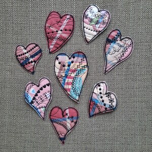 May include: Seven handmade fabric hearts in various shades of pink and red with black stitching. The hearts are made from different patterned fabrics and are all unique.