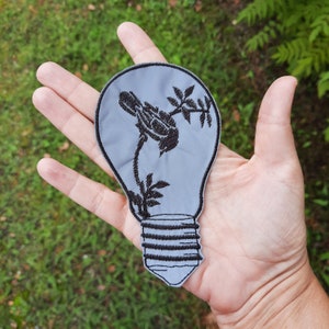 Może przedstawiać: Szara łatka w kształcie żarówki z czarnym haftem stylizowanej rośliny z kwiatami i liśćmi. Łatka znajduje się na dłoni.
