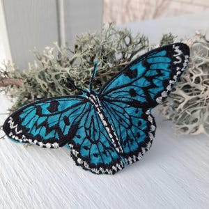 Puede incluir: Una mariposa azul turquesa con detalles en blanco y negro, bordada en tela. La mariposa se muestra sobre una superficie blanca con un fondo verde claro.