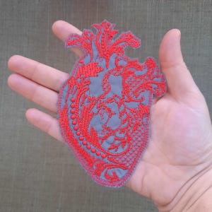May include: A red embroidered heart-shaped patch with a detailed floral design. The patch is on a gray background.