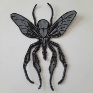 Op de afbeelding: Zwarte geborduurde strijkpatch van een gestileerd insect met vleugels. Het insect heeft een grijs lichaam en zwarte poten.