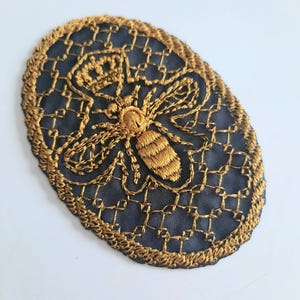 May include: A gold embroidered bee design on a black mesh fabric oval patch. The bee is detailed with a crown and wings.