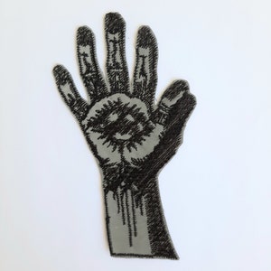 May include: Black and white embroidered patch of a hand with an eye in the palm.