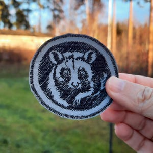 May include: A round, embroidered patch featuring a black and white illustration of a raccoon's face. The patch has a light gray background and a black, zigzag stitched border. The design is detailed and appears to be hand-stitched.
