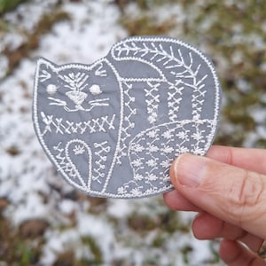 May include: A grey cat-shaped patch with detailed white embroidery. The patch depicts a cat with floral and cross-stitch patterns. The cat's body is grey, and the embroidery is white.