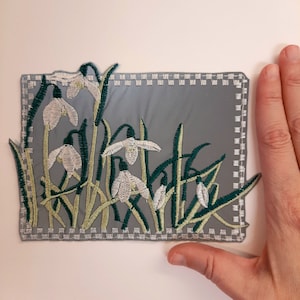Peut inclure: Un patch en feutre gris avec une bordure blanche cousue, représentant un motif brodé de perce-neige blancs avec des feuilles vertes.