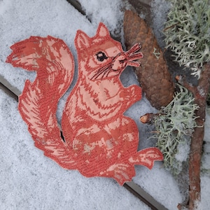 May include: A red embroidered squirrel patch with a detailed design. The patch is on a white snowy surface.