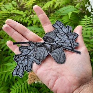 Op de afbeelding: Een grijs geborduurd patch van een eikenblad met twee eikels. Het eikenblad is gedetailleerd met nerven en de eikels zijn getextureerd.