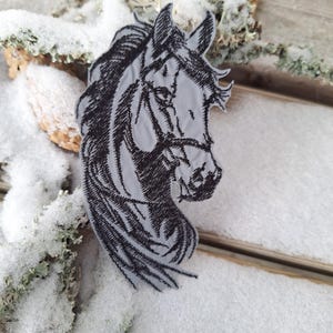 Peut inclure: Un écusson de tête de cheval grise avec des détails brodés noirs. L'écusson présente un contour détaillé de la tête et de la crinière d'un cheval. L'écusson est posé sur un fond enneigé et rustique.