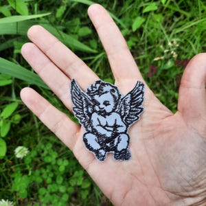 May include: A grey embroidered iron-on patch of a cherub angel with black wings. The angel is sitting with its hands clasped in front of it.