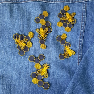 Könnte beinhalten: Fünf gestickte Aufnäher mit Bienen und Waben auf einer blauen Jeansjacke. Die Aufnäher sind gelb und schwarz und in einem zufälligen Muster angeordnet.