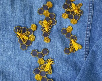 Peut inclure: Cinq patchs brodés représentant des abeilles et des rayons de miel sur une veste en jean bleue. Les patchs sont jaunes et noirs et sont disposés de manière aléatoire.