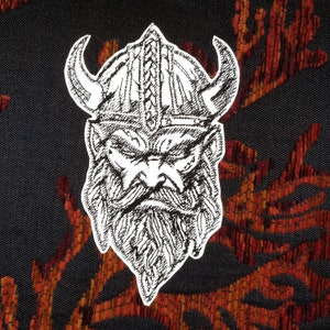 May include: A black and white embroidered patch of a Viking warrior wearing a horned helmet with a beard. The patch is on a red and black fabric background.