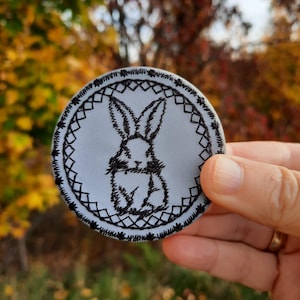 Puede incluir: Un parche redondo bordado de color gris con un contorno negro. El parche presenta un conejo negro estilizado con orejas largas dentro de un marco circular.