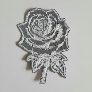May include: A white embroidered rose patch on a grey background. The rose is detailed with intricate stitching and has a stem and leaves.