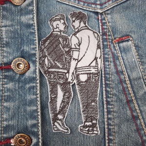 May include: A brown embroidered patch of two people standing side by side. The patch is sewn onto a blue denim jacket.