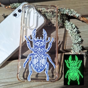 Op de afbeelding: Een doorzichtig telefoonhoesje met een blauw geborduurd vliegend hert-ontwerp. De keverafbeelding is centraal op de achterkant van het hoesje geplaatst. Een inzetstuk toont hetzelfde keverontwerp dat fel neongroen oplicht in een donkere omgeving.