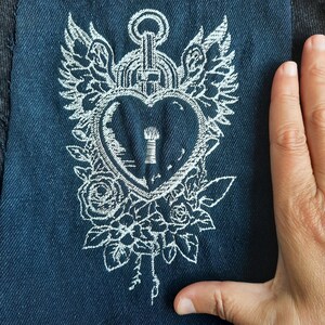 Puede incluir: Diseño bordado en blanco sobre una tela vaquera azul oscuro. El diseño presenta un corazón con un ojo de cerradura, rodeado de alas y flores.