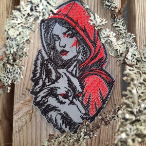 Peut inclure: Patch brodé représentant une femme dans une cape à capuche rouge, tenant un loup. Le visage de la femme est visible et la tête du loup est tournée vers le spectateur. Le patch est sur un fond en bois.