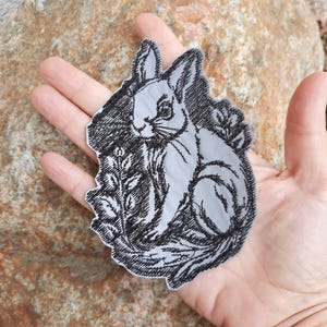 May include: A black and white embroidered iron-on patch featuring a rabbit sitting in a field of flowers.