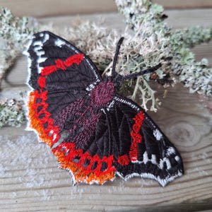 May include: Embroidered butterfly patch with a detailed design. The butterfly features black wings with red, orange, and white accents. The body is a deep purple colour. The patch is likely for clothing or accessories.