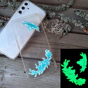 May include: A clear smartphone case featuring two turquoise embroidered patches with a jagged, organic shape. The patches are designed to glow in the dark, as shown in the inset image where they emit a bright green light against a black background.