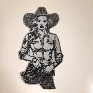 Può includere: Toppa ricamata in bianco e nero di una donna con un cappello da cowboy e una camicia abbottonata. Tiene una pistola nella mano destra.