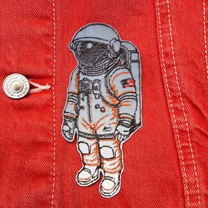 Puede incluir: Parche bordado de un astronauta con un traje espacial blanco con detalles naranjas. El astronauta lleva un casco y una mochila. El parche está en una chaqueta vaquera roja.