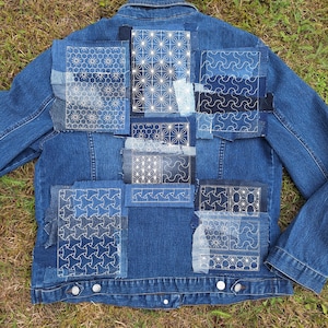 May include: A denim jacket with patches of various sizes and shapes. The patches are adorned with white embroidered geometric patterns. The jacket has a classic collar, button closures, and two chest pockets. The jacket is a medium wash blue.