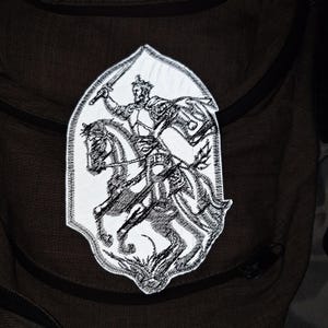 Pode incluir: Um patch bordado branco com uma ilustração a preto e branco de um cavaleiro a cavalo, brandindo uma espada. O patch tem uma borda decorativa e está sobre um fundo escuro.