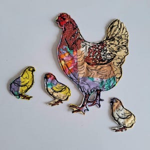 Puede incluir: Un conjunto de apliques de tela con una gallina y tres pollitos. La gallina es un patchwork de telas coloridas, con una cresta roja y costuras detalladas. Los pollitos son amarillos, morados y marrones, con contornos negros. Ideal para proyectos de costura.