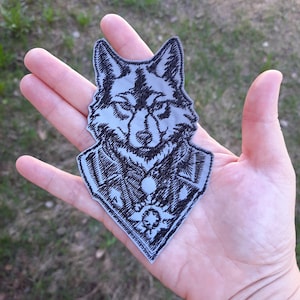 May include: A gray embroidered patch of a wolf's head with a detailed design. The wolf is wearing a collar with a decorative element.