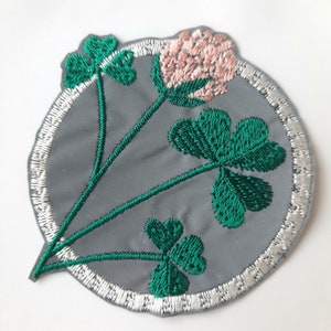Puede incluir: Un parche de trébol verde bordado con una flor rosa en el centro, rodeado de un círculo blanco sobre un fondo gris.