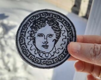 May include: Black and white embroidered patch featuring a portrait of a woman with a gear design around her head. The patch has a white background and black stitching.