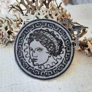 Könnte beinhalten: Ein grauer gestickter Aufnäher mit einem Schwarzweißbild eines Frauen Gesichts. Der Aufnäher ist rund und hat einen schwarzen Rand mit Text um den Rand herum.