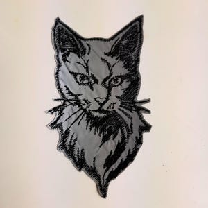 Pode incluir: Patch bordado de cabeça de gato em preto e branco. O gato está olhando diretamente para o espectador com uma expressão séria. O patch é feito de um tecido cinza com bordado preto.