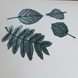 Può includere: Quattro foglie di tessuto ricamate in tonalità di verde e grigio. Le foglie sono disposte su uno sfondo bianco.