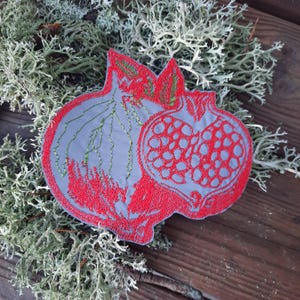 May include: A red embroidered pomegranate design on a gray fabric background. The pomegranate is detailed with green leaves and seeds.
